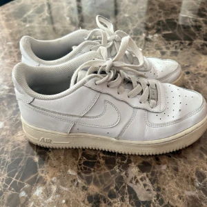 Vita Nike Air Force 1 sneakers - Klassiska vita Nike Air Force 1 sneakers i skinn med perforerad tå, snörning och låg profil. Snygg och tidlös modell med vit sula och diskret Nike-logga på sidan. Perfekta för en clean och sportig stil.