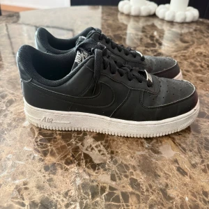 Svarta Nike Air Force 1 sneakers - Säljer ett par klassiska svarta Nike Air Force 1 sneakers med vit sula. Skorna har snörning, rund tå och är tillverkade i skinn med textilfoder. Perfekta för dig som gillar streetstyle och vill ha en tidlös look.