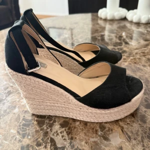 Svarta kilklackar med öppen tå - Snygga svarta sandaler med hög kilklack i flätad jute och öppen tå. Skorna har en justerbar rem med spänne runt ankeln och ovandel i mockaliknande material. Perfekta för sommarens alla outfits.