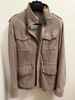 Rose & Born jacka - Stilren mockajacka från Rose & Born i beige med fyra stora fickor framtill och axelklaffar. Jackan har klassisk krage, tryckknappar och mjukt grått innerfoder. Perfekt för dig som vill ha en tidlös och snygg look.