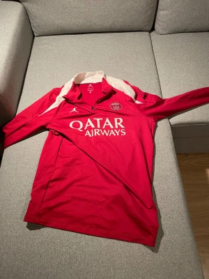 PSG långärmad röd fotbollströja Jordan - Paris Saint-Germain långärmad fotbollströja i rött med vita detaljer och dragkedja vid halsen. Tillverkad i Dri-Fit-material som andas. PSG-logga och Jumpman-logo på bröstet samt Qatar Airways-tryck framtill. Perfekt för dig som älskar fotboll och vill sticka ut.