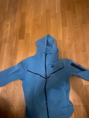 Blå hoodie från Nike med dragkedja - Säljer en blå hoodie från Nike med hel dragkedja framtill och svart Nike-logga på bröstet. Tröjan har luva , svarta detaljer längs dragkedjan och en ficka med dragkedja på ena ärmen. Perfekt för chill eller träning.