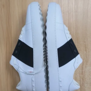 Valentino Garavani vita sneakers - Säljer ett par snygga vita Valentino Garavani sneakers med bred svart rem över ovansidan. Skorna har snörning, platt sula med coola pyramidformade detaljer och rund tå. Tillverkade i skinn för en lyxig känsla och stilren look.