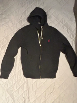 Svart hoodie från Polo Ralph Lauren - Svart hoodie från Polo Ralph Lauren med dragkedja och vit snörning i huvan. Klassisk röd Polo-logga på bröstet. Långärmad och med ribbade muddar. Perfekt för chill dagar och enkel att matcha med jeans eller joggers.