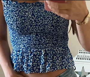 Blommig blå linnetopp från Pigalle - Supersöt blå linnetopp från Pigalle med vitt blommigt mönster. Toppen har smockad, tight passform och volangkant nedtill. Axelbanden är breda och rynkade för extra detalj. Perfekt för varma dagar och ger en fräsch vibe till din outfit.