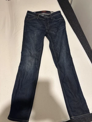 Mörkblå jeans från Jack & Jones 29/32 - Snygga mörkblå jeans från Jack & Jones med klassisk femficksdesign och kontrastsömmar. Modellen är Regular Clark och har en rak passform som funkar till det mesta. Tillverkade i slitstarkt denimtyg för en tidlös look.