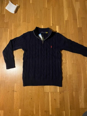 Mörkblå kabelstickad tröja Polo Ralph Lauren - Snygg mörkblå kabelstickad tröja från Polo Ralph Lauren med half zip och klassisk röd broderad logga på bröstet. Tröjan är gjord i 100% bomull och har ribbad krage. Perfekt för dig som gillar stilrena och tidlösa plagg. 