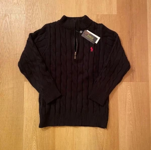Svart stickad tröja Polo Ralph Lauren - Svart kabelstickad tröja från Polo Ralph Lauren med halv dragkedja och röd broderad logga på bröstet. Tröjan har ribbade muddar och hög krage, perfekt för kyliga dagar. Klassisk och stilren design som funkar till många olika looks. Helt ny 