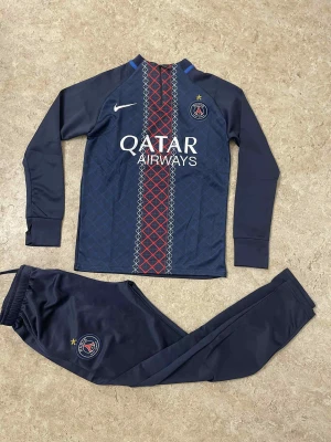 PSG långärmad fotbollströja Nike - Säljer en mörkblå långärmad fotbollströja från Nike med PSG-logga och Qatar Airways-tryck på bröstet. Tröjan har ett snyggt geometriskt mönster i rött och vitt längs mitten, samt matchande byxor med PSG-emblem. Materialet är lätt och andas, perfekt för träning.