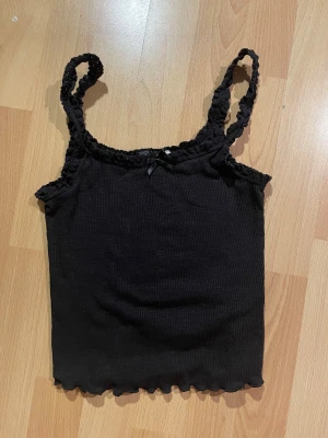 Svart ribbad linnetopp från H&M - Svart ribbad linnetopp från H&M med volangkant och liten rosett framtill. Axelbanden är smala och dekorerade med spetsdetaljer.