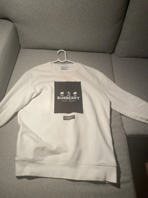 Vit sweatshirt från Burberry - Vit sweatshirt från Burberry med svart fyrkantigt tryck och logga på bröstet. Tröjan har rund hals, långa ärmar och är tillverkad i mjuk bomull. Snyggt tryck med globmotiv och texten 'London, England'. Perfekt för dig som gillar stilrena designer.