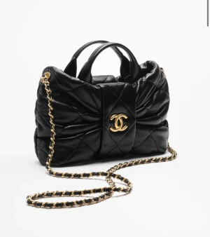 Svart quiltad handväska  - Supersnygg svart handväska med quiltad design och stor rosett framtill. Väskan har guldfärgad kedja som axelrem och ikonisk CC-logga i guld på framsidan. Tillverkad i mjukt skinn och har dragkedja inuti. Perfekt för dig som vill ha något lyxigt och unikt.