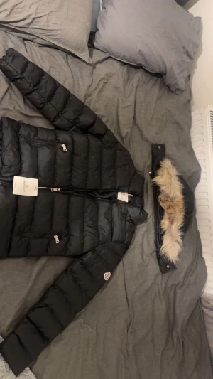 Svart Moncler dunjacka med pälskrage - Svart dunjacka från Moncler med avtagbar pälskrage. Jackan har klassisk quiltad design, dragkedjor på fickorna och Moncler-logga på ärmen. Perfekt för kalla dagar och riktigt snygg till streetwear-stilen.