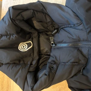 Svart pufferjacka Sail racing - Svart pufferjacka från Sail racing med broderad logga på bröstet och patch på ärmen. Jackan har dragkedja framtill, justerbar huva och kardborre vid ärmslut. Perfekt för kalla dagar och riktigt snygg streetstil. Pris kan diskuteras vid snabb affär 