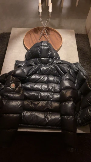 Svart Moncler pufferjacka med huva - Säljer en svart pufferjacka från Moncler med glansig finish och stor huva. Jackan har Moncler-logga på ärmen, breda quiltade sektioner och dragkedja. Insidan har en cool serietidningsetikett. Perfekt för dig som vill ha en fet vinterjacka med street vibe. Pris kan diskuteras vid snabb affär‼️