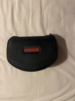 Zonefloorball sportglasögon med extra linser - Säljer ett par sportiga Zonefloorball glasögon med svart båge och tre utbytbara linser: klara, mörka och spegel. Allt kommer i ett robust svart fodral med dragkedja och Zonefloorball-logga. Perfekta för innebandy eller annan sport där du vill skydda ögonen.