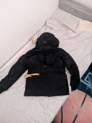 Svart Parajumpers parkajacka med huva - Svart parkajacka från Parajumpers med flera fickor, huva och snygga detaljer i orange och gult. Jackan har dragkedja, knappar och patch på ärmen. Perfekt för kalla dagar med sin fodrade insida och robusta känsla.