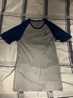 Grå och marinblå Gymshark t-shirt - Snygg t-shirt från Gymshark i storlek S med grå kropp och marinblå raglanärmar. T-shirten har rund hals och diskret Gymshark-logga på bröstet. Perfekt för träning eller chill, tillverkad i mjuk polyesterblandning som andas.