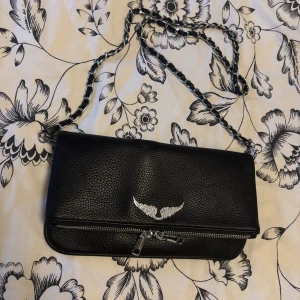 Svart axelväska med kedja och vingar - Snygg svart axelväska i skinn med silverfärgad kedja som axelrem. Väskan har ett dekorativt emblem med glittriga vingar på framsidan och dragkedja både fram och bak. Perfekt storlek för det viktigaste och cool detalj till din outfit.