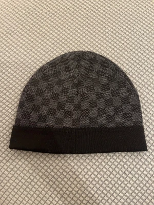  svart rutig mössa  - Svart mössa  med klassiskt rutigt mönster och flera färgglada LV-patchar framtill. Mössan har uppvikt kant och är tillverkad i ett mjukt material som passar perfekt för kyliga dagar. Snygg accessoar med exklusiv känsla och streetstyle-vibe.