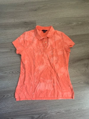 Korall pikétröja Tommy Hilfiger - Snygg korall orange/rosa pikétröja från Tommy Hilfiger med klassisk krage och tre knappar framtill. Tröjan har ett unikt blekt mönster och den ikoniska Hilfiger-loggan broderad på bröstet. Perfekt för dig som gillar färg och vill sticka ut lite extra.