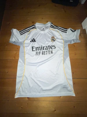 Real Madrid hemmatröja Adidas vit - Säljer en Real Madrid hemmatröja från Adidas. Tröjan är vit med svarta detaljer och gula linjer längs sidorna. Klassisk klubbmärke på bröstet, Adidas-logga och 'Emirates Fly Better' tryck på framsidan. Tillverkad i lätt funktionsmaterial. PRIS KAN DISKUTERAS!!!!!
