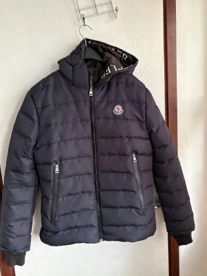 Svart Moncler pufferjacka S - Snygg svart pufferjacka från Moncler i storlek S. Jackan har horisontella sömmar, två dragkedjefickor och en hög krage med Moncler-logga på insidan. Klassisk Moncler-patch på bröstet och innerfoder med serietidningsdetalj. Perfekt för kalla dagar.