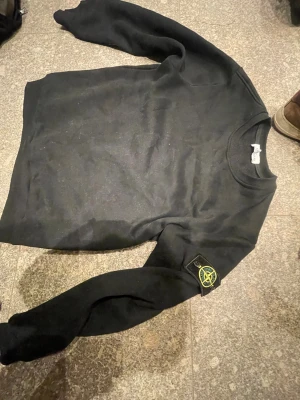 Svart Stone Island sweatshirt  - Svart sweatshirt från Stone Island i storlek M. Klassisk rund hals och långärmad modell med den ikoniska Stone Island-loggan på vänster ärm. Tröjan är tillverkad i mjuk bomull och har en clean, stilren och shyst look till alla olika outfits. Bara att höra av sig vid funderingar