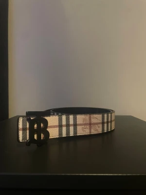 Rutig bälte med spänne - Snyggt bälte med klassiskt rutigt mönster i beige, svart och rött. Bältet har ett stilrent svart BB-spänne framtill och är gjort i skinn. Perfekt accessoar för att lyfta din outfit.