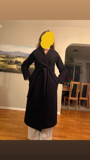 Svart lång kappa med bälte - Säljer en svart, lång kappa med bred krage och bälte i midjan. Klassisk omlottstil med knappar och fickor på sidorna. Kappan har en stilren och tidlös look, perfekt för dig som gillar minimalistisk vibe och vill hålla dig varm under höst och vinter. Finns en liten defekt och det är att knappen inuti för att atänga inifrån fattas dock väldigt enkelt att sy dit en ny! Från bubbleroom nypris 1100kr!