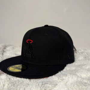 Svart New Era 59FIFTY LA Angels keps - Snygg svart LA Angels keps från New Era 59FIFTY med röd broderad logga och detaljer. Platt skärm och klassisk passform, perfekt för dig som gillar streetwear och sport. Materialet är bomull och kepsen har broderade ventilationshål. Kan vara lite mindre i storlek 