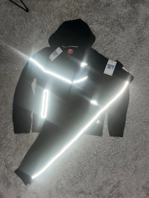 Svarta Nike suit med reflexdetaljer - Säljer ett par svarta mjukisbyxor från Nike med vita reflexdetaljer längs benen och logga på låret. Byxorna har ribbade muddar vid bensluten och är gjorda i mjukt material som är perfekt för chill eller träning. Snygg och modern design med sportig känsla.