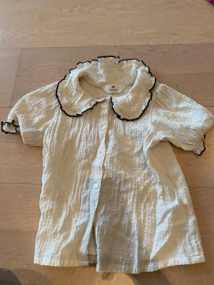 Vit blus med volangkrage från H&M - Supersöt vit blus från H&M i storlek 116. Den har korta puffärmar, volangkrage med svart kant och knappar framtill. Materialet är krinklat och känns luftigt och lätt. Perfekt för dig som gillar romantiska detaljer och en lekfull stil.