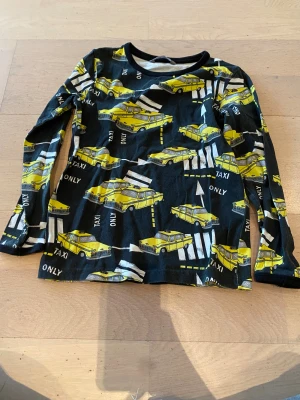 Svart långärmad taxi-tröja Lindex - Cool svart långärmad tröja från Lindex med tryck av gula taxibilar och vita vägmarkeringar över hela plagget. Rund hals och mjukt bomullsmaterial som är skönt mot huden. Perfekt för dig som gillar unika och färgglada prints.