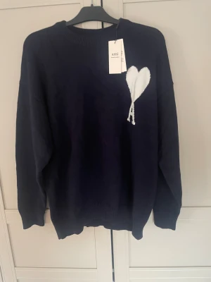 Ami tröja - Snygg mörkblå stickad tröja från Ami med vit hjärtapplikation och ett A på bröstet. Tröjan har rund hals, ribbade muddar och lång ärm. Perfekt för dig som gillar stilrena detaljer och vill ha något som sticker ut lite extra. Helt ny, passade inte mig 