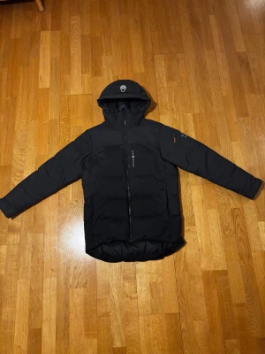 Sail Racing jacka - Svart dunjacka från Sail Racing, modell Antarctica Expedition. Jackan har huva, hel dragkedja framtill och diskreta loggor på bröstet och ärmen. Vadderad och lång i modellen, perfekt för kalla dagar. Snygg och stilren design med praktiska detaljer. Använd en säsong, ser helt ny ut. Om ni har några frågor komm pv!