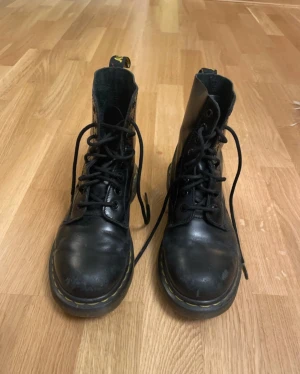 Svarta Dr. Martens Pascal boots - Säljer ett par svarta Dr. Martens Pascal boots i skinn med klassisk gul söm runt sulan. Modellen har snörning, rund tå och platt sula. Ikonisk dragflik baktill med Dr. Martens-logga. Perfekt för dig som gillar en edgy stil och vill ha boots som sticker ut.