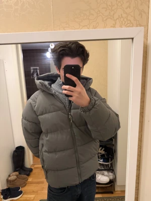 Grå pufferjacka från Pull&Bear - Säljer en grå pufferjacka från Pull&Bear i storlek S. Jackan har huva, dragkedja framtill och två sidofickor med dragkedja. Den är quiltad och har en loose passform som ger en chill vibe. Materialet är polyester och jackan är perfekt för kalla dagar.