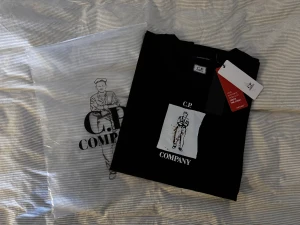Svart C.P. Company t-shirt med print - Svart t-shirt från C.P. Company med coolt grafiskt print framtill som visar en tecknad figur och logga. Klassisk passform och rund hals. Kommer med originalpåse och taggar kvar. Perfekt för dig som gillar streetwear och stilrena detaljer.