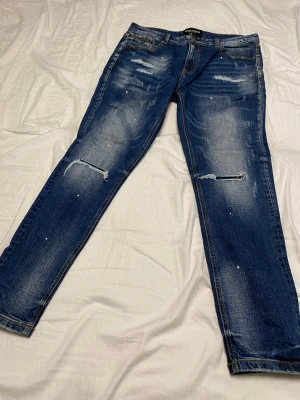 Blå Supply & Demand jeans  - Snygga blå jeans från Supply & Demand med slitna detaljer på benen. Jeansen har klassisk femficksdesign, dragkedja vid ena fickan och kontrastsömmar. 