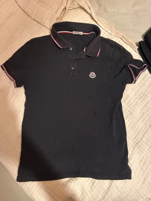 Mörkblå Moncler pikétröja med röd/vit kant - Svart pikétröja från Moncler med klassisk krage och knappar framtill. Tröjan har röda och vita detaljer på krage och ärmslut samt Moncler-logga broderad på bröstet. Tillverkad i bomull och har korta ärmar. Perfekt för en clean och sportig look.