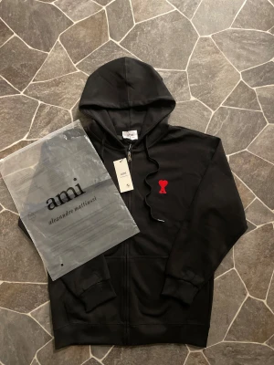 Svart hoodie från Ami Paris - Svart hoodie från Ami Alexandre Mattiussi med dragkedja och huva. På bröstet finns ett broderat rött hjärta med bokstaven A. Tröjan har ribbade muddar och snörning vid huvan. Materialet är mjuk bomull, perfekt för chill dagar. Passar både M och L.