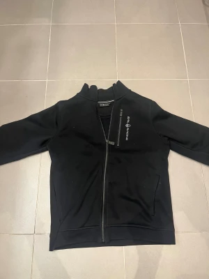 Svart hoodie från Sail Racing, strl 160 - Svart hoodie från Sail Racing med hög krage och tvåvägsdragkedja framtill. Jackan har en bröstficka med dragkedja och broderad logga på bröstet. Tillverkad i mjukt material som känns skönt mot huden. Perfekt för dig som gillar stilren och sportig look.