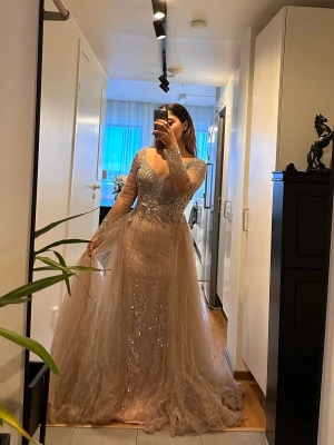 Champagnefärgad glittrig balklänning - Min förlovning fest klänning,köpte själv för 8000kr.Magisk långklänning i champagnefärg med glittriga pärldetaljer och broderier. Klänningen har v-ringning, långa transparenta ärmar och ett svepande tyllsläp. Materialet är lätt och skimrande, perfekt för bal eller fest. Snygg passform som markerar midjan och ger en lyxig känsla.