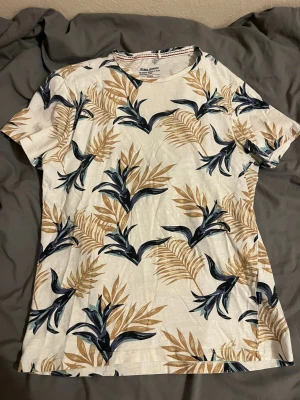 Vit t-shirt med bladmönster från Blend - Säljer en vit t-shirt från Blend med ett tropiskt bladmönster. T-shirten har korta ärmar och är perfekt för en avslappnad stil.