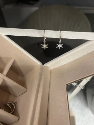 Silvriga örhängen med stjärnhänge - Säljer ett par snygga silvriga örhängen med hängande stjärnor. Örhängena har en minimalistisk design och är perfekta för dig som gillar detaljer med lite edge. Stjärnorna är små och blanka, vilket ger ett stilrent och coolt intryck.