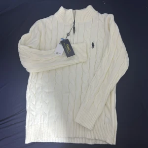 Vit kabelstickad tröja Polo Ralph Lauren - Kabelstickad vit tröja från Polo Ralph Lauren med halv dragkedja och broderad logga på bröstet. Tröjan har ribbade muddar och hög krage. Kom dm för pris 