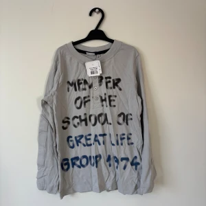 WeKids långärmad tröja - 122/128 Storlek - Grå långärmad tröja från WeKids med tryckt text i svart och blått framtill. Rund halsringning och knappslå med tre knappar. Tillverkad i mjuk bomull. 