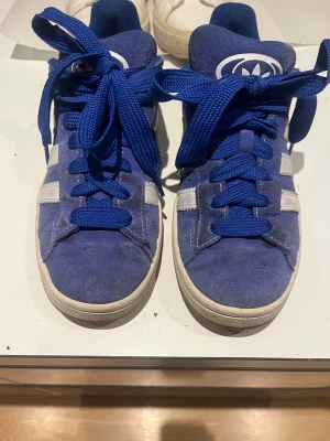 Blå Adidas Campus sneakers i mocka - Säljer ett par blå Adidas Campus sneakers med klassiska vita ränder på sidorna och vit sula. Skorna har rund tå, platt sula och är gjorda i mocka med blå snörning. Perfekta för dig som gillar retrostil och vill sticka ut med färg på fötterna.