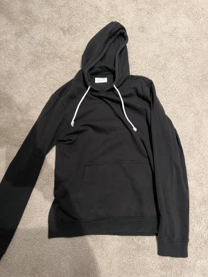 Svart hoodie från Pier One - Svart hoodie från Pier One med klassisk huva och vita snören. Tröjan har en stor magficka framtill och är långärmad. Perfekt för chill dagar och enkel att matcha med jeans eller joggers. Materialet känns mjukt och skönt mot huden.